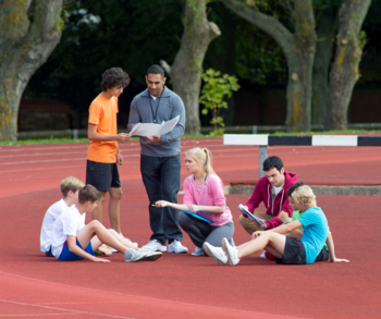 Gruppe junger Menschen auf einem Sportplatz mit ihrem Trainer: 5 sitzen auf dem Boden, ein Kind spricht mit dem Trainer